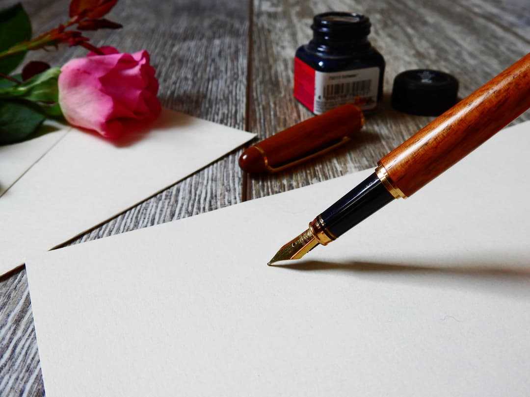 a piece of paper on a wooden table with an ink pen held over it, about to write a letter. A rose petal, the pen cap and an opened bottle of ink sit next to it.