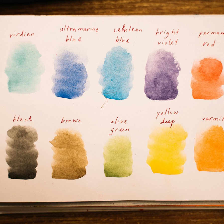 Ten brush strokes on white paper of a watercolour palette arranged horizontally across two lines, with five colours on each line. Each colour is labeled above its respective stroke with its name to identify each hue.