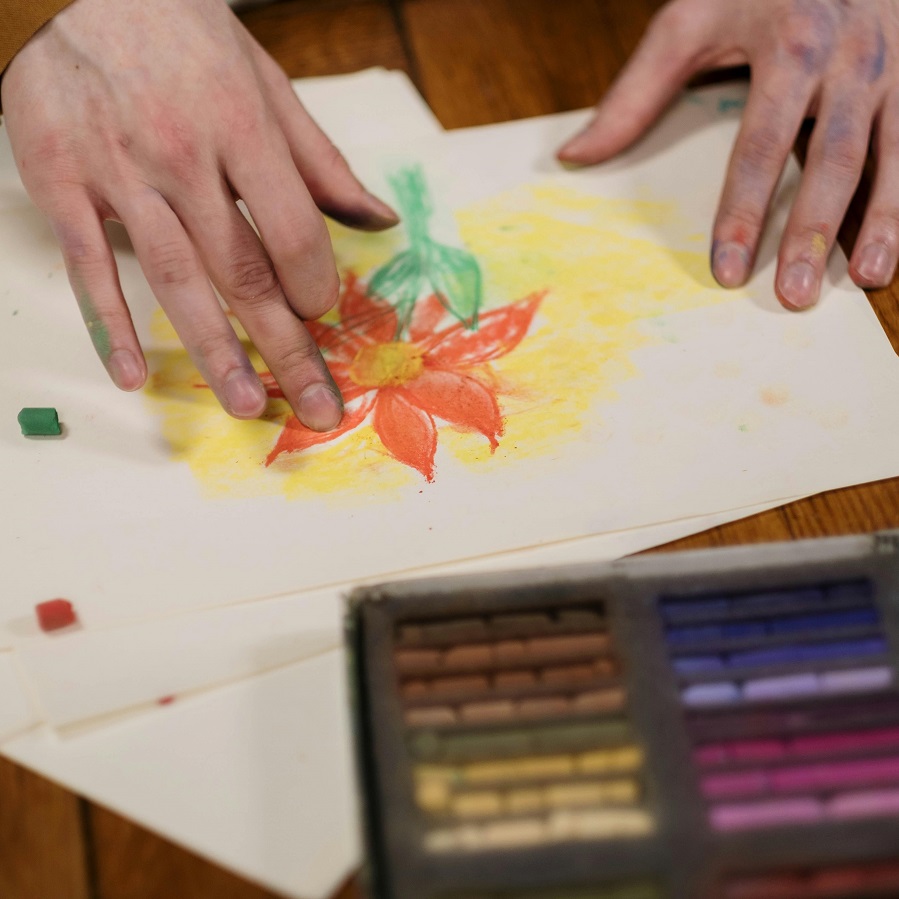 Hands over a pastel drawing of an orange flower. A box of pastels lies next to it.